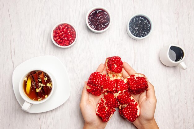 Top view plate on the white table peeled pomegranate in hands bowls of jam and sauce and a cup of herbal tea with lemon on the table