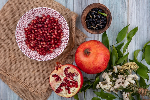 Top view of pomegranate berries in bowl on sackcloth with whole and half ones and bowl of blackberry with flowers on wooden surface