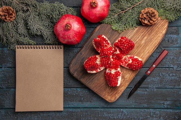 Free photo top view pomegranate on board pilled pomegranate on cutting board next to spruce branches with cones knife and cream notebook on grey surface