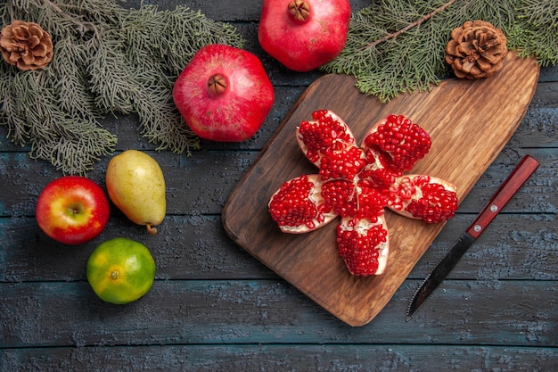 Free photo top view pomegranate on board pilled pomegranate on cutting board next to spruce branches with cones knife lime apple pear on grey surface