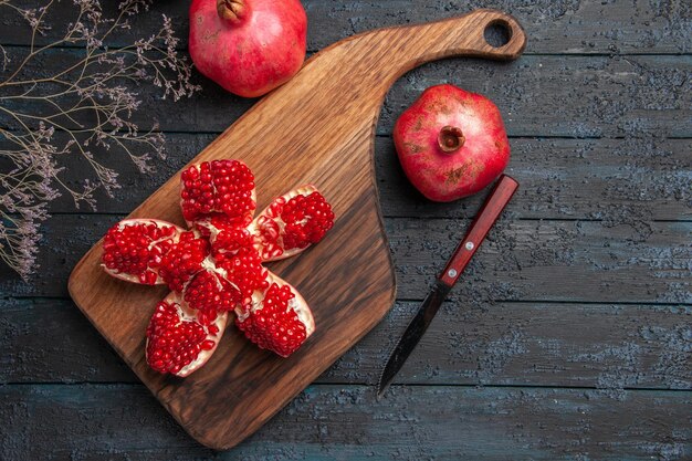 Top view pomegranate on board pilled pomegranate on cutting board next to tree branches knife between two pomegranates on dark table