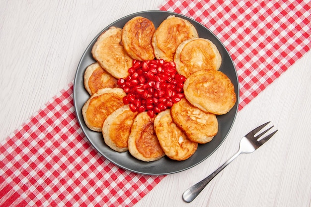 Top view pomegranate pancakes and seeds of pomegranate on the checkered tablecloth and a fork on the table