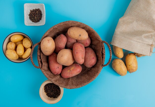 Free photo top view of potatoes in basket and in bowl with other ones spilling out of sack and black pepper seeds on blue