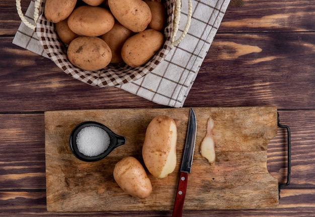 Top view of potatoes with shell knife and salt on cutting board with other ones in basket on cloth on wooden surface