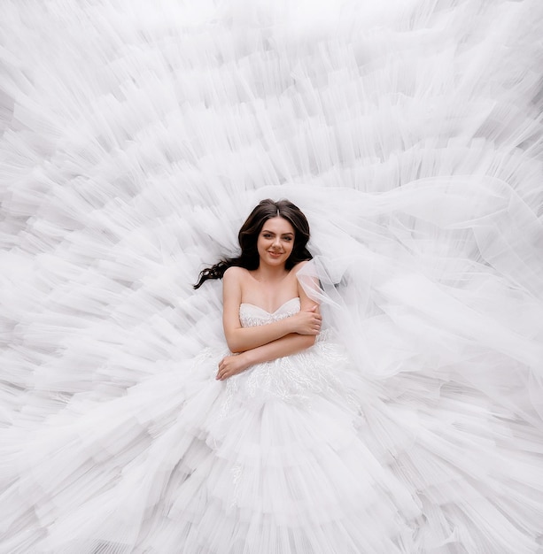 Free photo top view of pretty brunette model bride girl lying on puffy white wedding dress with open shoulders elegant lady dressed in princess style dress