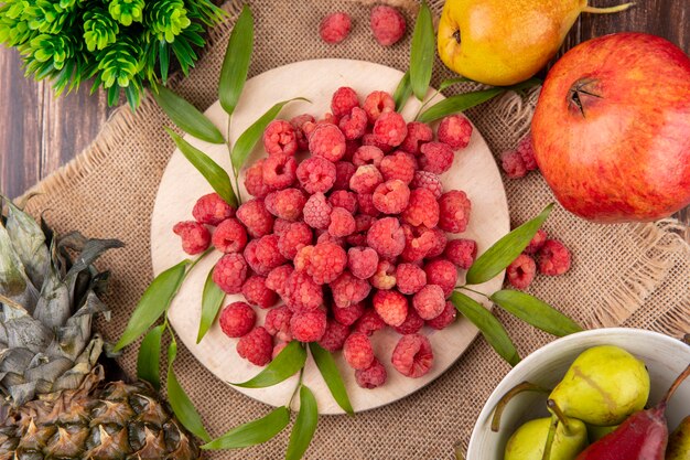 Top view of raspberries with leaves on cutting board and pineapple and pomegranates on sackcloth and wooden surface