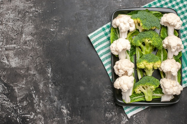 Top view raw broccoli and cauliflower on black rectangular plate on green and white checkered napkin fork and knife on dark surface free space