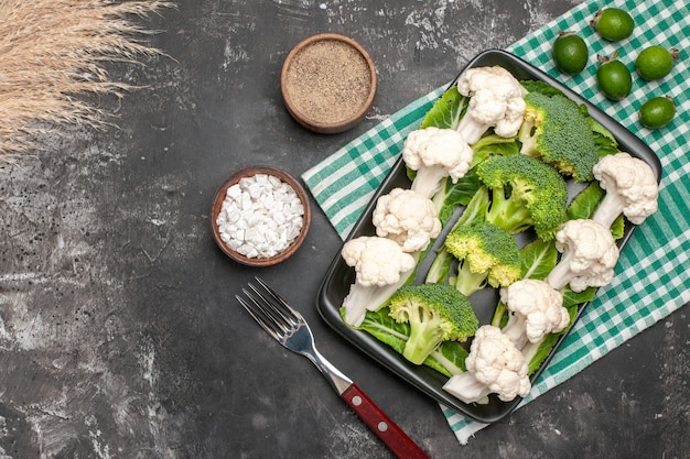 Free photo top view raw broccoli and cauliflower on black rectangular plate on green and white checkered napkin fork sea salt pepper feykhoas on dark surface free space