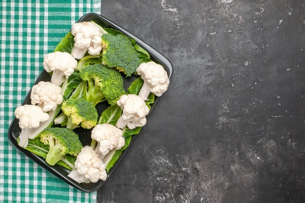 Top view raw broccoli and cauliflower on black rectangular plate on green and white checkered tablecloth on dark surface free place