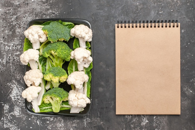 Free photo top view raw broccoli and cauliflower on black rectangular plate a notebook on dark surface