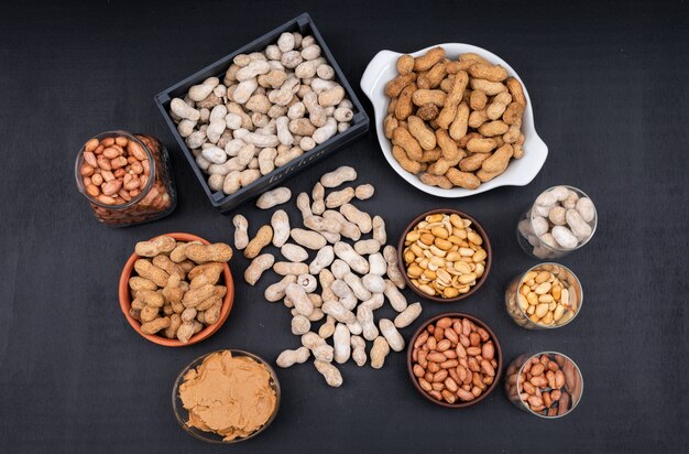 Top view raw, fresh, fried peanuts and peanut butter in box, jar and bowl, on dark 