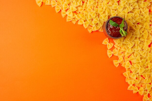 Top view of raw pasta farfalle with ketchup in a saucepan and copy space on orange