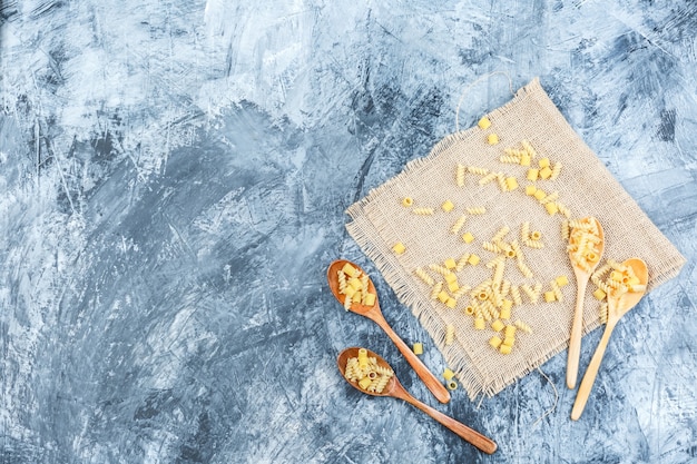 Free Photo top view raw pasta in wooden spoons on plaster and piece of sack background. horizontal