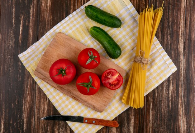 Top view of raw spaghetti with tomatoes on a cutting board with a knife and cucumbers on a yellow checkered towel on a wooden surface