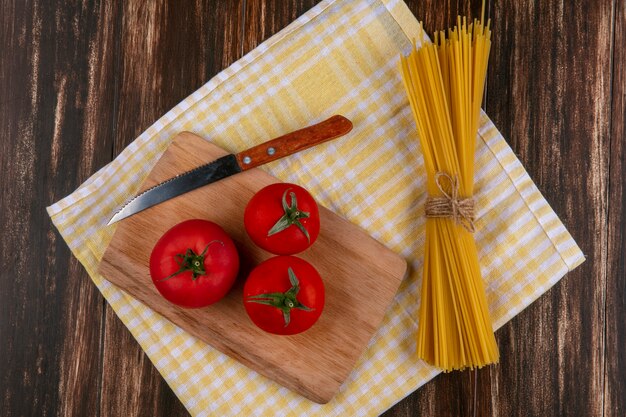 Top view of raw spaghetti with tomatoes on a cutting board with a knife on a yellow checkered towel on a wooden surface