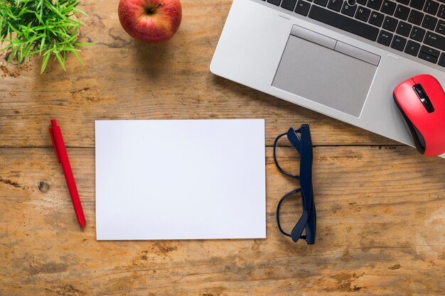 Top view of red apple; mouse; laptop; pen; eyeglasses and blank white paper on wooden desk