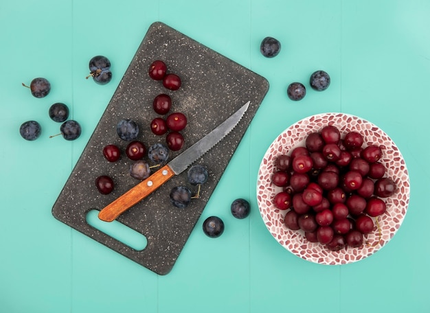 Free photo top view of red cherries on a bowl with sloes on a kitchen cutting board with knife on a blue background