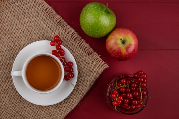 Top view red currant in a glass with a cup of tea and apples on a red background