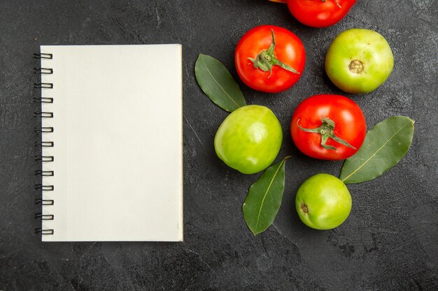 Top view red and green tomatoes bay leaves and a notebook on dark surface