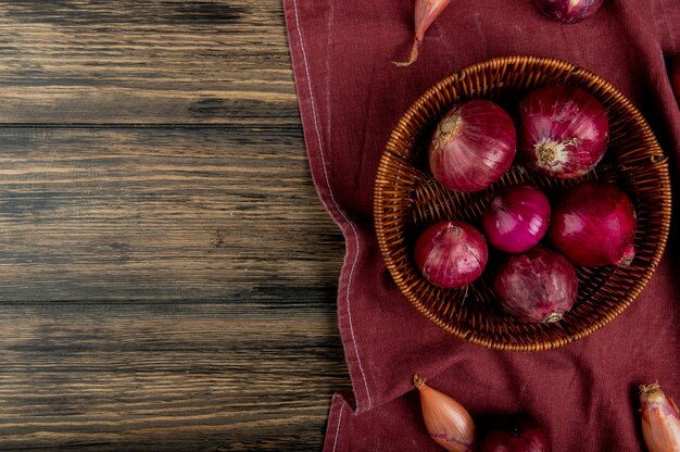 Top view of red onions in basket with shallots on bordo cloth and wooden background with copy space