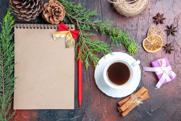 Top view red pen a notebook pine tree branches xmas tree ball toys and gifts a cup of tea white saucer cinnamon sticks anises on dark red surface