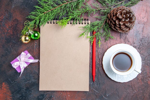 Top view red pen a notebook pine tree branches xmas tree toys and gifts a cup of tea on dark red surface