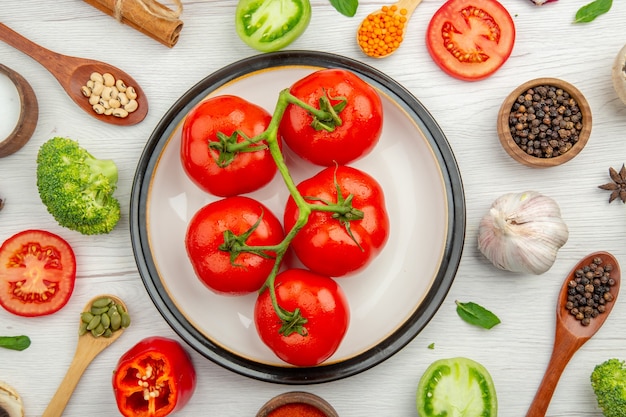 Top view red tomatoes on plate black pepper in bowl wooden spoons garlic broccoli on grey table