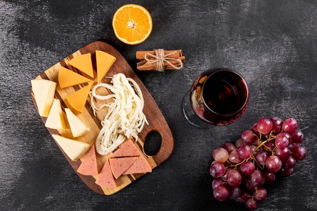 Top view of red wine with grape, orange and cheese on wooden cutting board on dark surface horizontal