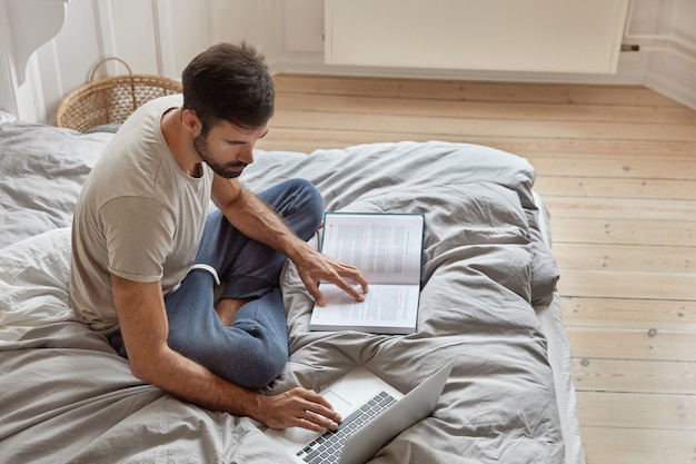 Top view of relaxed bearded guy poses at cozy bed in lotus pose, ponders on readed material, checks information from book in laptop computer, studies laws, works in bedroom. Domestic atmosphere