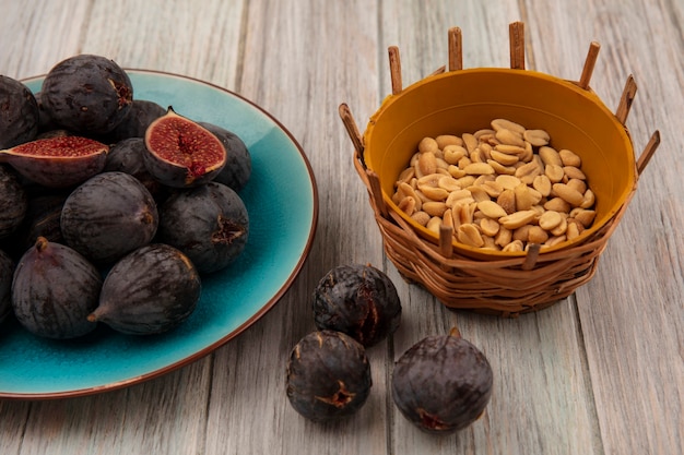 Top view of ripe black mission figs on a blue bowl with peanuts on a bucket on a grey wooden wall