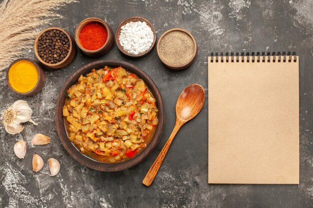 Top view of roasted eggplant salad in bowl different spices in bowls garlic notebook on dark surface