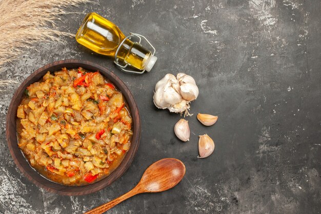 Top view of roasted eggplant salad in bowl oil bottle garlic wooden spoon on dark surface