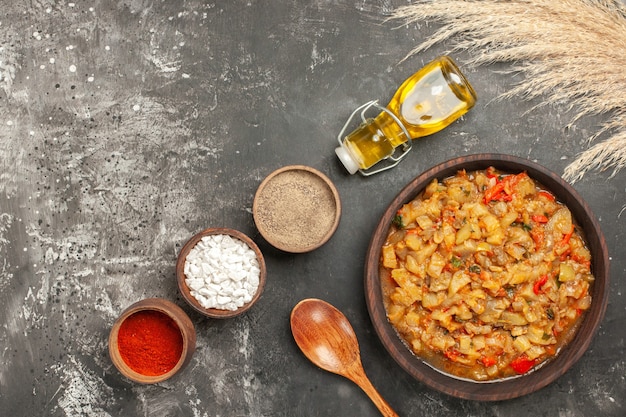 Top view of roasted eggplant salad in bowl, oil bottle, wooden spoon and different spices in bowls on dark surface