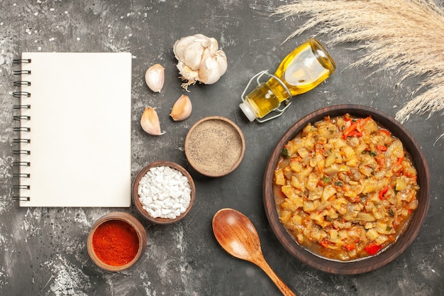 Free photo top view of roasted eggplant salad in bowl, oil bottle, wooden spoon and different spices in bowls notebook on dark surface