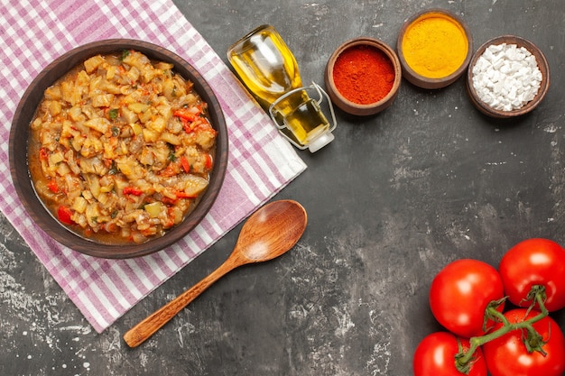 Free photo top view of roasted eggplant salad in bowl oil, wooden spoon and tomatoes on dark surface