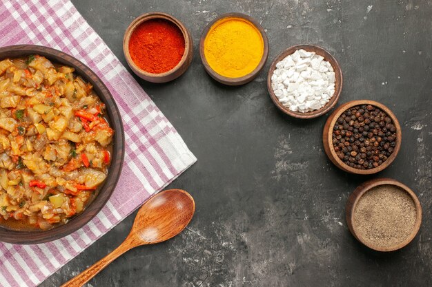 Top view of roasted eggplant salad in bowl wooden spoon different spices in bowls on dark surface