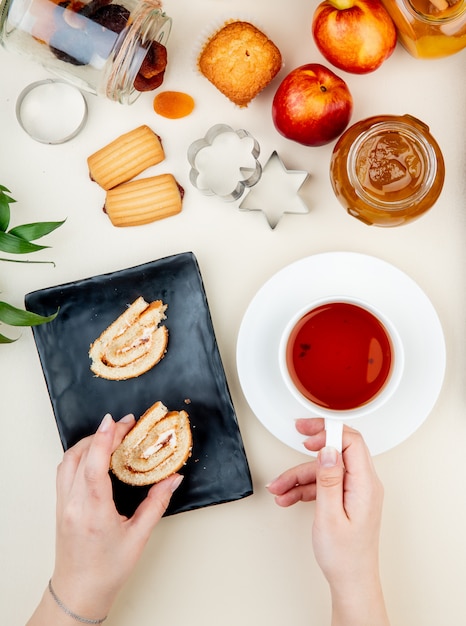 top view of roll cake on a black tray served with a cup of tea and a glass jar with peach jam cookies fresh ripe nectarines and cookie cutters on white