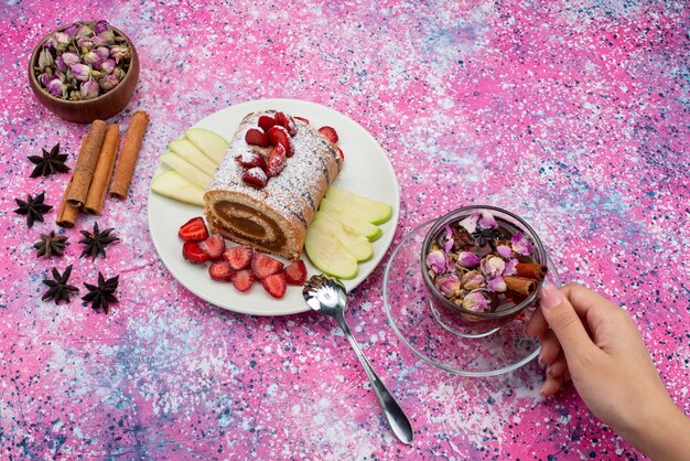 Top view roll cake inside plate with apples and strawberries along with cinnamon and tea on the colored desk cake biscuit sweet fruit