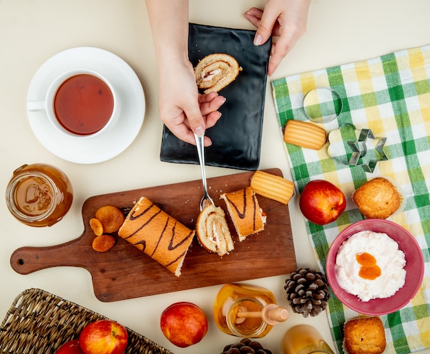 Top view of roll cake laying on a black tray and holding a cup of tea and a glass jar with peach jam cookies fresh ripe nectarines  cottage cheese and cookie cutters on white backgro