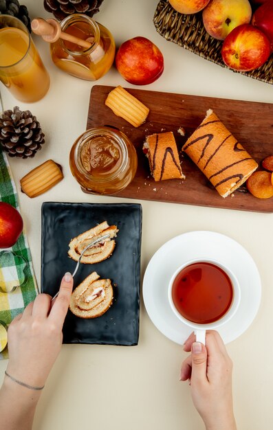 Top view of roll cake laying on a black tray and holding a cup of tea and a glass jar with peach jam cookies fresh ripe nectarines and a glass of juice on white