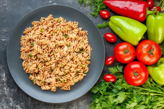 Top view rotini pasta on round plate bell peppers tomatoes parsley dill cherry tomatoes on grey table