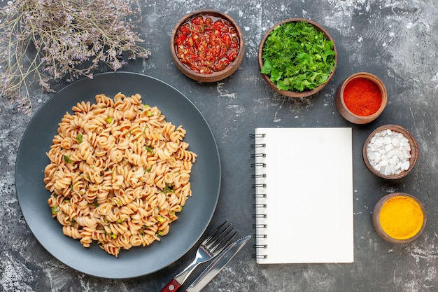 Top view rotini pasta on round plate fork and knife sea salt turmeric red pepper powder in small bowls notepad on grey table