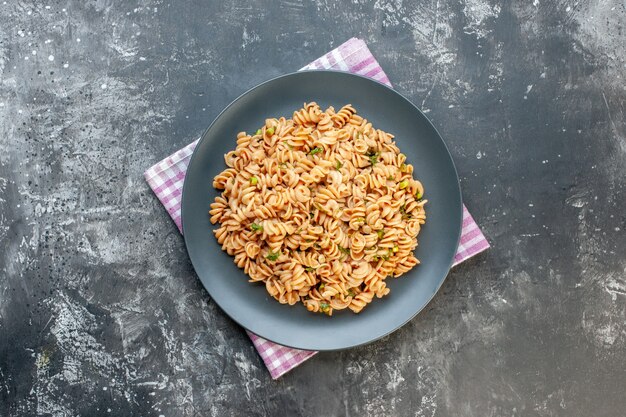 Top view rotini pasta on round plate on pink white checkered kitchen towel on dark surface copy place