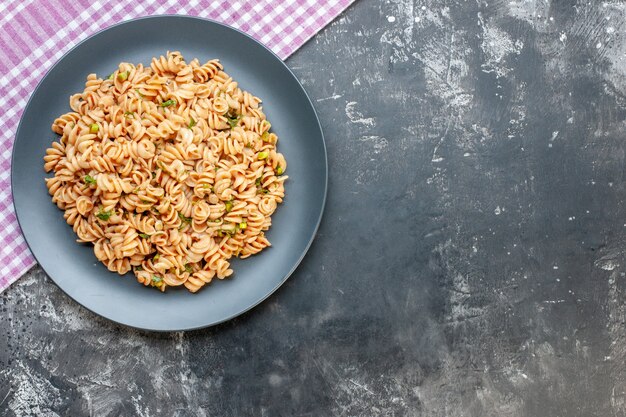 Top view rotini pasta on round plate on pink white checkered tablecloth on dark surface with copy space