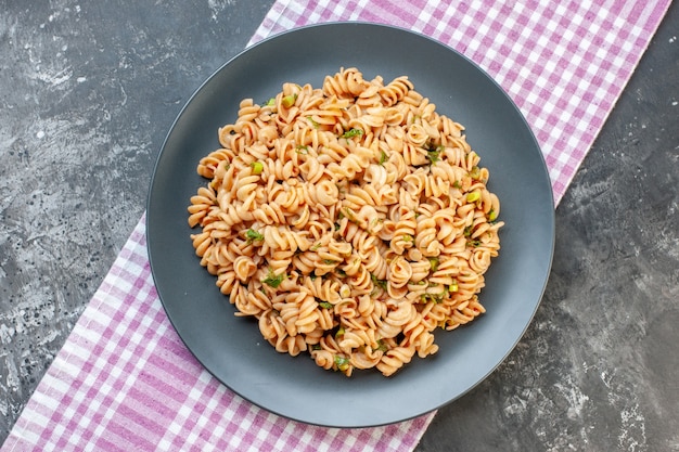 Top view rotini pasta on round plate on pink white checkered tablecloth on dark surface