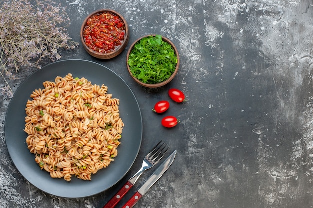 Top view rotini pasta on round plate tomato sauce chopped parsley in small bowls cherry tomatotes fork and knife on dark table free space