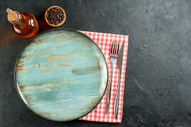 Free Photo top view round platter dinner knife and fork black pepper in small bowl oil bottle red and white checkered napkin on black table