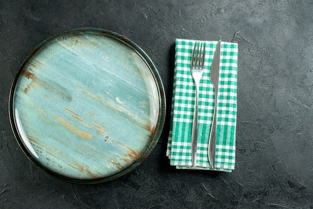 Top view round platter dinner knife and fork on green and white checkered napkin on black table free space