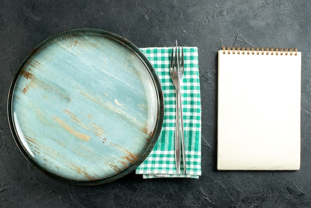 Top view round platter dinner knife and fork on green and white checkered napkin notebook on black table