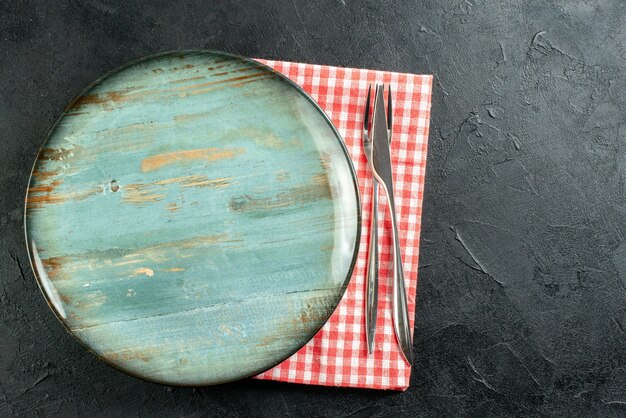 Top view round platter dinner knife and fork on red and white checkered napkin on black table free space